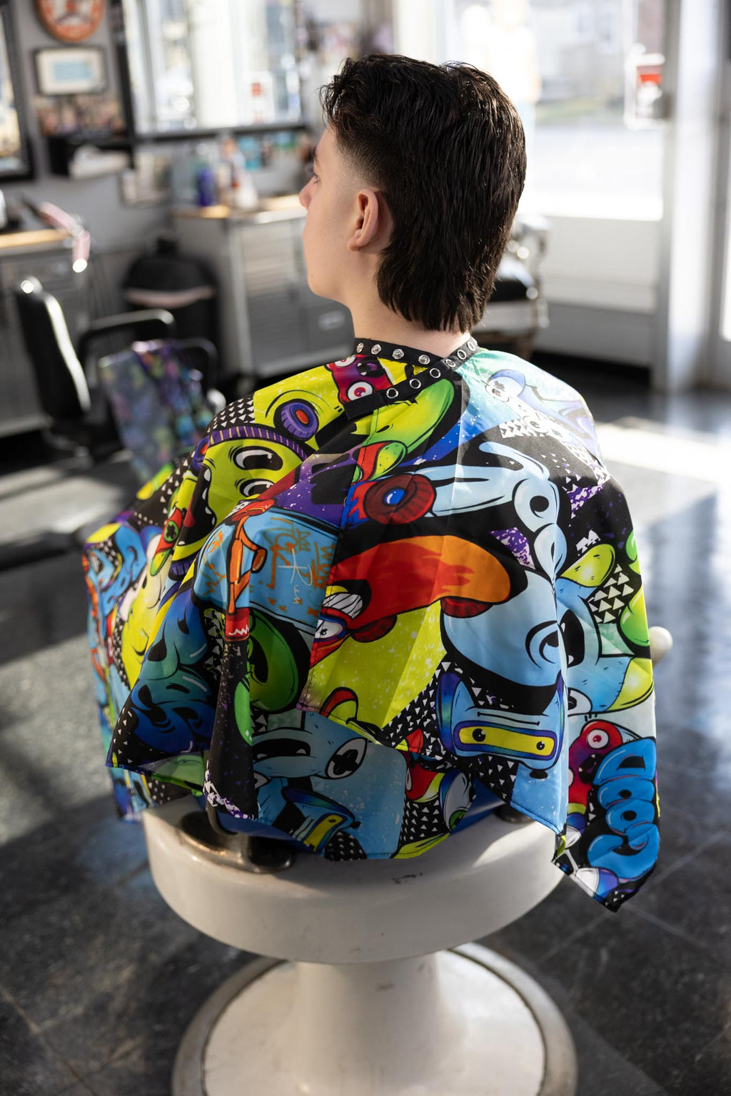 Graffiti Themed waterproof professional barber and salon cape with metal snap closures and unique design including the vibrant colorful style featuring graffiti style gaming controllers skateboards and words play and cool
