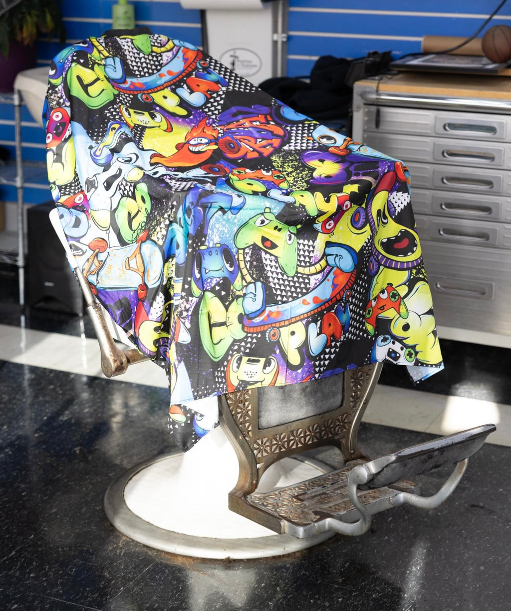 Graffiti Themed waterproof professional barber and salon cape with metal snap closures and unique design including the vibrant colorful style featuring graffiti style gaming controllers skateboards and words play and cool